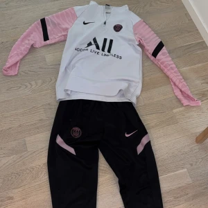 PSG långärmad fotbollströja från Nike - Säljer en långärmad PSG fotbollströja från Nike. Tröjan är vit med rosa ärmar och svarta detaljer, inklusive klubbmärke och sponsortryck på bröstet. Perfekt för träning eller match. Matchande svarta byxor med rosa detaljer ingår.
