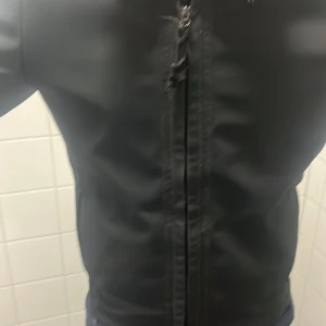 Svart jacka från Stone Island med dragkedja - Säljer en svart jacka från Stone Island med klassisk logotyp på ärmen. Jackan har dragkedja framtill och en stilren, minimalistisk design. Perfekt för dig som gillar streetwear och vill ha något snyggt och enkelt.