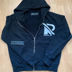 Replay Hoodie  - Svart hoodie från Replay med stor vit logga och text på både fram- och baksida. Tröjan har huva, dragkedja och muddar vid ärmslut och nederkant. Tillverkad i bomull och passar perfekt för en avslappnad stil. OPS hoodie har ett litet hål vid slutet av armen, som går enkelt att sy ihop. För mer frågor skriv gärna 