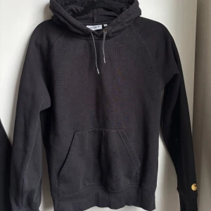 Svart hoodie från Carhartt - Säljer en svart hoodie från Carhartt med klassisk huva och dragsko. Tröjan har en stor magficka och Carhartt-logga i gult på ärmen. Perfekt för en avslappnad stil och enkel att matcha med det mesta.