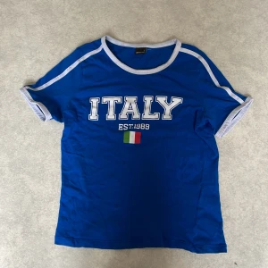 Blå t-shirt med ITALY-tryck från Gina Tricot - Blå t-shirt från Gina Tricot med vit kant vid hals och ärmar. Framsidan har stort vitt ITALY-tryck, texten EST.1989 och en liten italiensk flagga. Klassisk passform och kortärmad modell, perfekt för dig som gillar sportig stil.