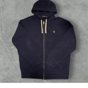 Mörkblå hoodie från Polo Ralph Lauren - Säljer en mörkblå hoodie från Polo Ralph Lauren med dragkedja och huva. Tröjan har en gul broderad logga på bröstet och vita snören vid huvan. Perfekt för en avslappnad stil.