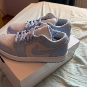 Nike Air Jordan 1 Low i ljusblå och vit - Säljer ett par fräscha Nike Air Jordan 1 Low i en snygg ljusblå och vit färgkombination. Skorna har klassisk snörning, rund tå och är tillverkade i skinn med syntetdetaljer. Perfekta för dig som gillar stilrena sneakers med ikonisk design.