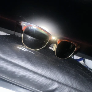 Clubmaster solglasögon från Ray-Ban - Snygga Clubmaster solglasögon från Ray-Ban med en klassisk design. Bågarna är i en brun nyans med guldiga detaljer och mörka linser. Perfekta för en stilren look.