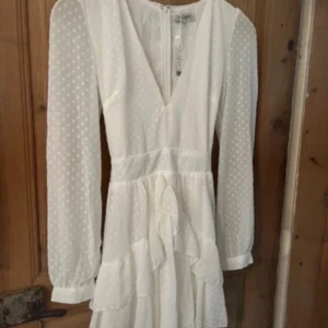 Student klänning - Superfin vit kortklänning från Showpo med diskret prickigt mönster, v-ringning och volangdetaljer. Klänningen har långa ärmar, knytband i midjan och dragkedja i ryggen. Perfekt för sommarens tillfällen!