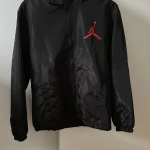 Svart Jordan vindjacka  - Svart vindjacka från Jordan med röd Jumpman-logga på bröstet och stor logga samt texten 'JORDAN' på ryggen. Jackan har huva, dragkedja och resår vid ärmslut och midja. Perfekt för sportiga dagar. Helt oanvänd då den var för liten för mig. 