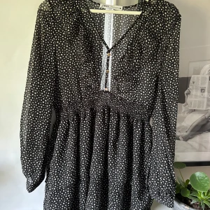 Svart prickig klänning från Zara - Säljer en svart klänning från Zara med vita prickar och långa ärmar. Klänningen har v-ringning med knappar framtill och smock i midjan för en fin passform.🌟