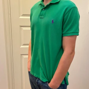  Ralph Lauren piké - Säljer en klassisk grön pikétröja från Polo Ralph Lauren med lila broderad logga på bröstet. Tröjan har korta ärmar, krage och knappar vid halsen. Perfekt för en stilren och avslappnad look. Modellen är 183 cm och pikén är i fint skick!