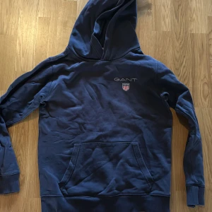 Mörkblå hoodie från GANT - Säljer en mörkblå hoodie från GANT med broderad logotyp och sköld på bröstet. Tröjan har huva, långa ärmar och en stor magficka framtill. 