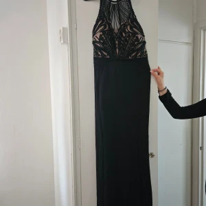 Svart långklänning med broderad mesh - Säljer en svart långklänning med broderad mesh upptill och hög halsringning. Klänningen har en snygg slits framtill och dragkedja i ryggen. Detaljerad brodyr i svart över beige bas ger en lyxig känsla. 