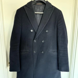 Mörkblå rock från Oscar Jacobson - Stilren mörkblå rock från Oscar Jacobson med dubbelknäppning och klassisk krage. Kappan har långa ärmar och två fickor framtill. Perfekt för dig som vill ha en tidlös och elegant look.