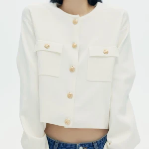 Vit croppad kofta guldknappar - Snygg vit croppad kofta med dekorativa guldknappar framtill och på ficklocken. Koftan har långa ärmar och två stora bröstfickor. Perfekt för en trendig och stilren look. Finatickat material.