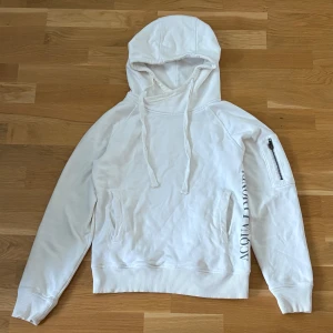 Acqua Limone Hoodie - Säljer en vit hoodie från Acqua Limone med klassisk citronlogga på ryggen och tryck längs sidan. Tröjan har huva med dragsko, fickor fram och en extra ficka med dragkedja på ärmen. Perfekt för en avslappnad stil. Sitter som xs/s.
