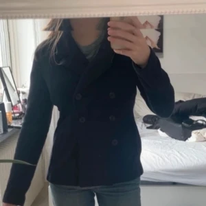Svart skepparkavaj - Säljer en klassisk svart skepparkavaj med dubbelknäppning och krage. Jackan har en figurnära passform och är perfekt för dig som gillar en stilren look. Passar till mycket och är enkel att matcha med olika outfits. 1 knapp saknas men är inget man tänker på och kan enkelt fixas med en ny knapp.