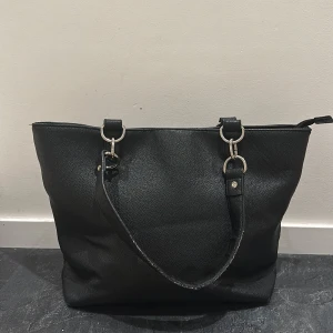 Svart Don Donna  axelväska med dragkedja - Säljer en svart Don Donna axelväska med silverfärgade metalldetaljer och dragkedja upptill. Väskan har en extra ficka med dragkedja på baksidan och tre fickor på insidan! 