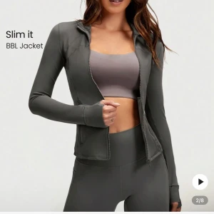 Mörkgrå träningsjacka med dragkedja Glowmode  - Slimmad mörkgrå träningsjacka med dragkedja och långa ärmar. Jackan har tumhål, liten dold ficka och sitter tight. Perfekt för träning eller sportiga outfits. Hip length-modell med minimalistisk design.