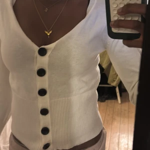 Vit kofta med svarta rosetter och stora knappar - Supersöt vit kofta med stora svarta knappar framtill och dekorativa svarta rosetter längs ärmen. Croppad modell med ribbad midja och ärmslut. Perfekt för dig som vill ha något unikt och stilrent🩷🩷