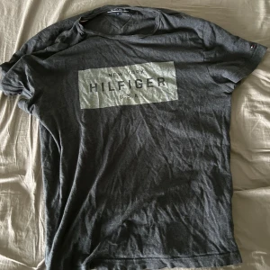 Mörkgrå t-shirt från Tommy Hilfiger - Säljer en mörkgrå t-shirt från Tommy Hilfiger med tryck på bröstet där det står 'New York Hilfiger U.S.A'. Klassisk passform och rund halsringning. Perfekt för en avslappnad stil.