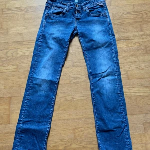 Blå jeansbyxor från Replay - Säljer ett par klassiska blå jeansbyxor från Replay med raka ben och normal passform. Byxorna har fem fickor, knappar i gylfen och tydliga slitningar för en cool look. Perfekta till vardags och lätta att matcha med olika stilar.