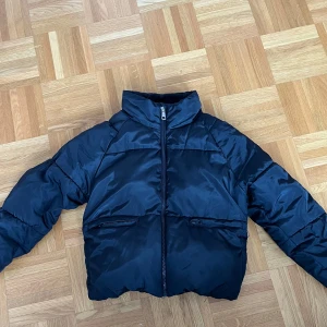 Svart pufferjacka från Lindex  - Svart pufferjacka från Lindex Kids med hög krage och dragkedja framtill. Jackan har två fickor med dragkedja och reflexdetaljer på ärm och rygg. Vattenavvisande material, perfekt för kyliga dagar.