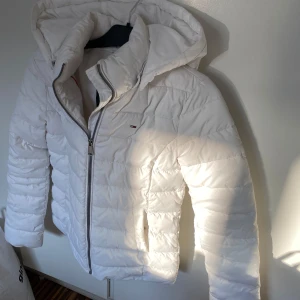 Vit pufferjacka från Tommy Jeans - Säljer en vit pufferjacka från Tommy Jeans med huva och dragkedja. Jackan har quiltad design och är perfekt för kyligare dagar. Klassisk logga på bröstet och innerkrage i mjukt material.