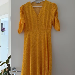 Ny Gul prickig klänning från Ellos. 38 - •Säljer en gul klänning från Ellos med vita prickar och v-ringning.  •Klänningen har knappar framtill, trekvartsärm med lätt puff och är i en mjuk, luftig kvalitet. •Den har en smickrande passform med en markerad midja och en utsvängd kjol.  •Klänningen har en dold dragkedja i sidan. 95 cm lång •Perfekt för sommarens alla tillfällen! •Ny utan lappar eftersom den är tvättad •Djurfritt och rökfritt hem