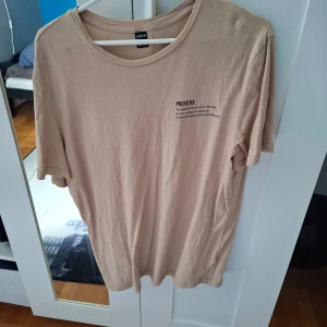 Beige t-shirt med tryck från SHEIN - Beige t-shirt från SHEIN i storlek M med rund hals och kort ärm. På bröstet finns ett svart tryck med texten 'PROVERB' och ett inspirerande citat. Enkel och stilren design, perfekt till vardags.