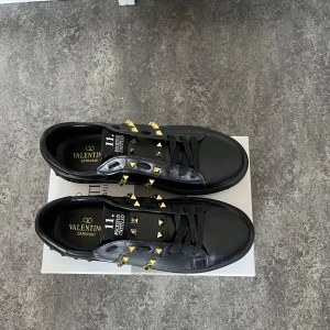 Svarta Valentino sneakers - Säljer ett par svarta Valentino Garavani sneakers med guldiga pyramidformade nitar längs sidorna. Skorna har snörning och platt sula, tillverkade i skinn. Snygg och edgy design som sticker ut.