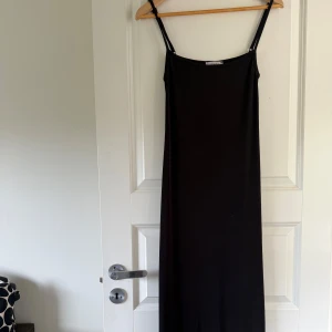 Svart långklänning med smala axelband - En stilren svart långklänning med smala axelband och rak passform. Perfekt för dig som gillar minimalistisk stil och vill ha en enkel men elegant klänning. Passar till många olika tillfällen.