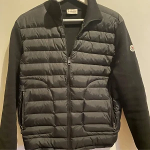  Moncler cardigan  - Säljer en svart pufferjacka från Moncler med quiltad front och ärmar i stickad bomull. Jackan har dragkedja framtill, två fickor med dragkedja och Moncler-logga på ärmen. Perfekt för kyliga dagar. Skick 9/10 lite defekt finns man ser knappast 