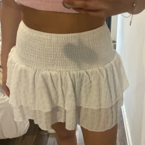 Vit volangkjol med smock i midjan - Säljer en söt vit kjol med smockad midja och dubbla volanger. Kjolen är kort och har en luftig känsla, perfekt för varma dagar. Passar dig som gillar en feminin och trendig stil.