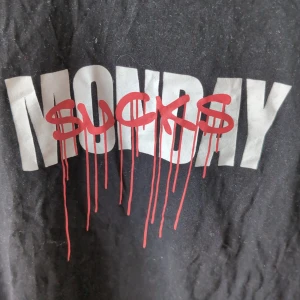 Svart t-shirt med tryck 'Monday Sucks' - Svart t-shirt  med stort vitt och rött tryck på bröstet där det står 'Monday Sucks' i graffiti-stil. Klassisk passform och rund halsringning. Perfekt för dig som vill göra ett statement.