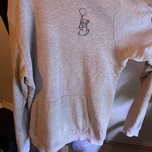 Grå hoodie med tryck från Brand Store - Grå hoodie från Brand Store med ett litet tryck av en tecknad figur som håller i en ballong på bröstet. Tröjan har magficka och huva. Perfekt för en avslappnad stil. Dock ett litet hål vid tröjans ärm, inget som inte går att sy.