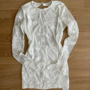 Vit långärmad miniklänning med blommiga spetsdetaljer - Jättefin vit långärmad miniklänning med blommiga spetsdetaljer och öppen rygg. Klänningen har tight passform och är perfekt för speciella tillfällen. Aldrig använt.