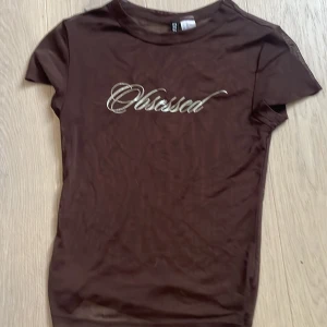 Brun mesh topp med texten 'Obsessed' från Divided - Säljer en brun mesh topp från Divided med glittrig text 'Obsessed' framtill. T-shirten är tight och har korta ärmar. Perfekt att styla lager på lager eller som statement-piece. Passar även S