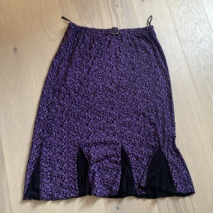 Lila blommig kjol med svarta detaljer - Säljer en lila kjol med små blommor och svarta inslag nertill. Kjolen har resår i midjan och en dekorativ detalj framtill. Perfekt för dig som gillar färg och mönster.står XL men passar mig som är M. Väldigt skönt material