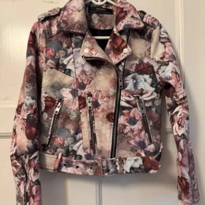 Blommig bikerjacka från Zara Basic - Säljer en snygg bikerjacka från Zara Basic med blommigt mönster i rosa, beige och grå toner. Jackan har dragkedjor,avtagbart bälte i midjan och klassisk krage.