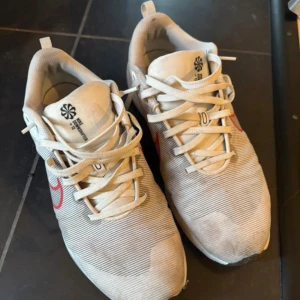 Vita Nike sneakers med röd swoosh - Säljer ett par vita Nike sneakers med röd swoosh på sidan. Skorna har snörning och en platt sula, samt diskreta ränder i materialet. Perfekta för träning eller vardagsstil. Klassisk design med bekväm passform.