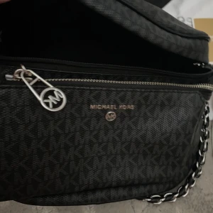 Svart axelväska från Michael Kors - Säljer en svart axelväska från Michael Kors med silverfärgad kedjedetalj och justerbart band. Väskan har dragkedja och logomönster över hela ytan samt en framficka. Perfekt för att bära dina viktigaste saker.