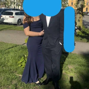 Marinblå långklänning med offshoulder - Elegant marinblå långklänning med offshoulder-design och draperad överdel. Klänningen har figurnära passform och lätt utsvängd kjol. Perfekt för bal eller fest. Tillverkad i ett slätt, följsamt material.