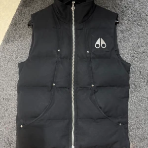Svart dunväst från Moose Knuckles - Säljer en svart dunväst från Moose Knuckles med dragkedja framtill och två fickor. Västen har en broderad logotyp på bröstet och ett broderat motiv på insidan. 