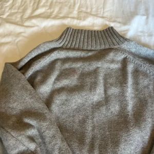 Grå stickad poloklänning i ullblandning - Säljer en mjuk och stilren grå polotröja med lång ärm och ribbade muddar. Tröjan har en hög polokrage och är perfekt för kyligare sommarkvällar eller höstväder. 