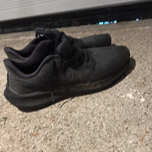 Svarta Nike sneakers Air Zoom - Säljer ett par helsvarta Nike Air Zoom sneakers med snörning och rund tå. Skorna har en sportig design med mesh-överdel och platt sula, perfekta för träning eller vardagsstil. Klassisk Nike-logga på sidan och bekväm passform. Nytt pris 1800 kr