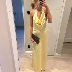 Ljusgul långklänning med öppen rygg och knyt - Säljer en ljusgul långklänning med draperad urringning och öppen rygg. Klänningen har knytband i nacken och är ärmlös, perfekt för sommarens tillfällen. Materialet är glansigt och ger en elegant känsla.