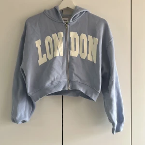 Ljusblå hoodie med LONDON-tryck från ZARA - Säljer en ljusblå hoodie från ZARA med vit LONDON-text framtill. Tröjan är croppad, har dragkedja och huva. Perfekt för en avslappnad stil och enkel att matcha med jeans eller leggings. Helt ny har änvänts ett fåtal gånger.