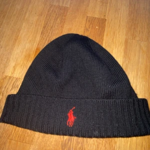 Svart mössa från Polo Ralph Lauren - Svart stickad mössa från Polo Ralph Lauren med röd broderad logga framtill. Tillverkad i mjuk och tvättbar merinoull, perfekt för kyliga dagar. Passar åldern 9-13 år. 
