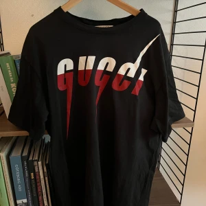 Svart t-shirt från Gucci med rödvit logga - Säljer en svart t-shirt från Gucci i modellen Blade. Stor, grafisk logga i vitt och rött framtill. Klassisk rund halsringning och korta ärmar. Tillverkad i mjuk bomull för en skön känsla. 