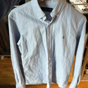 Ljusblå skjorta från Ralph Lauren - Klassisk ljusblå skjorta från Ralph Lauren med button-down krage och broderad logga på bröstet. Skjortan har långa ärmar och stängs med vita knappar framtill. Perfekt för en stilren och tidlös look.