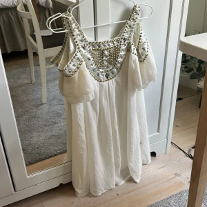 Vit klänning med pärlor och stenar - Supersöt vit klänning med axelbandslös look och glittriga detaljer. Överdelen är dekorerad med stora silvriga stenar och guldfärgade pärlor som ger en lyxig känsla. Klänningen har en luftig passform och faller fint, perfekt om du är ute i sista sekund för en studentklänning eller vill ha en i backup ;)