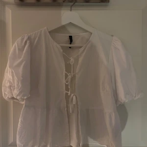 Vit blus med puffärmar från FB Sister - Säljer en söt vit blus från FB Sister med puffiga korta ärmar och knytning framtill. Blusen har en luftig passform och volang nedtill. Storlek M men mer som en S/XS